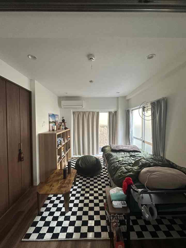 ベージュ色の壁と木製家具が調和した6畳の寝室、チェッカーボード柄のラグとビーズクッションが配された落ち着いた二人暮らしの空間