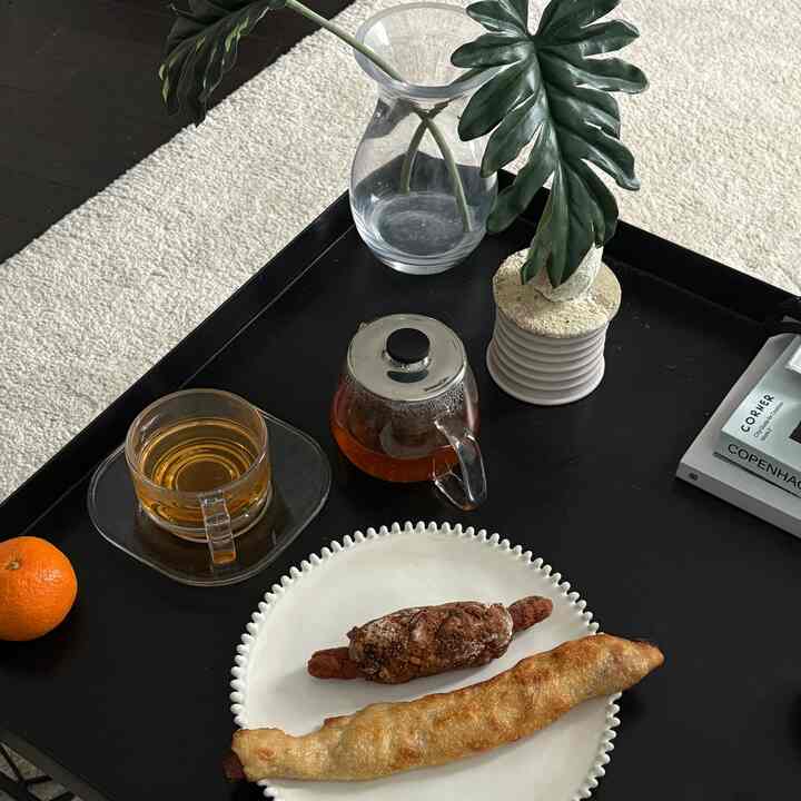 Natural modern living room with black tray coffee table on white rug, featuring a teacup, Kinto teapot, plant pot, and bread