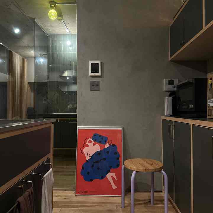 チャコールとダークグレーのキッチン空間、床に置かれた赤いポスターと薄紫の脚のスツールがあるモダンなインテリア