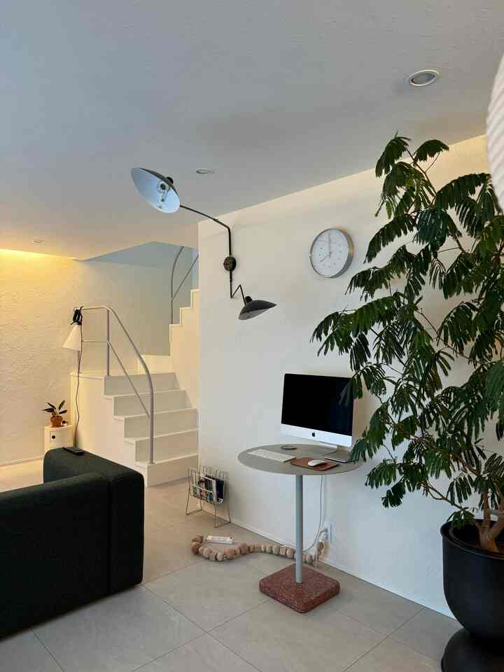 White-toned living room featuring dark green sofa, round table with computer, and natural plant, in Mid-Century Modern style