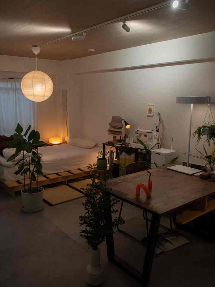Wood tone and white studio apartment featuring a bed and workspace with lighting and plants creating a cozy atmosphere