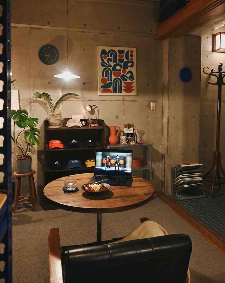 打ちっぱなしコンクリート壁のある小さなワンルーム、木製の丸テーブルと本棚、帽子が飾られた雰囲気のあるインテリア