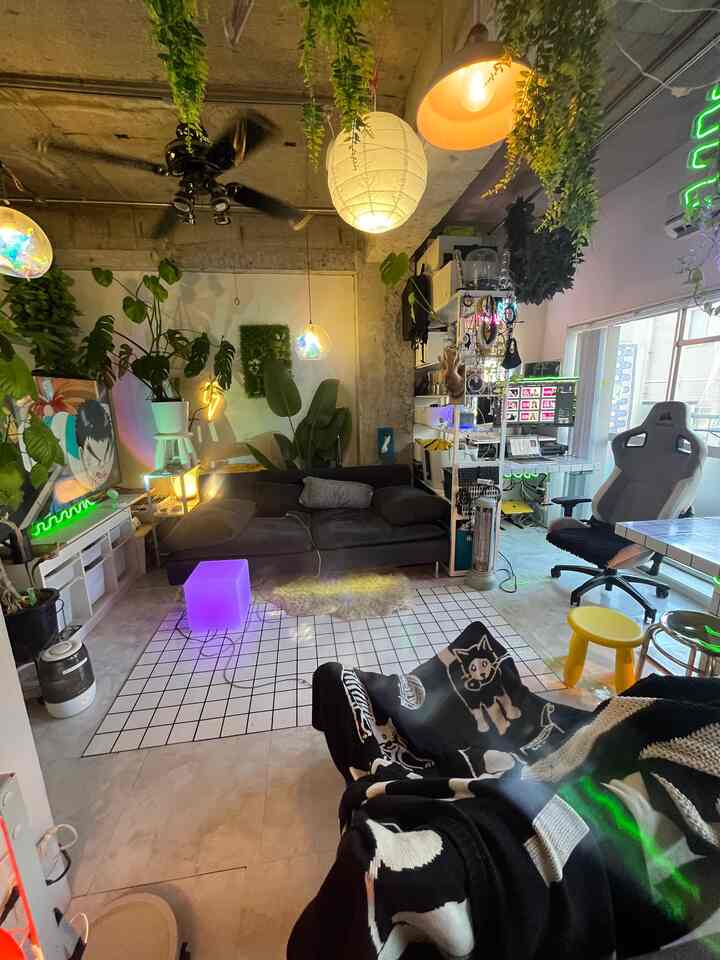 Industrial-style compact living room featuring a dark gray sofa, pendant lights, abundant plants, and a home office setup with a gaming chair