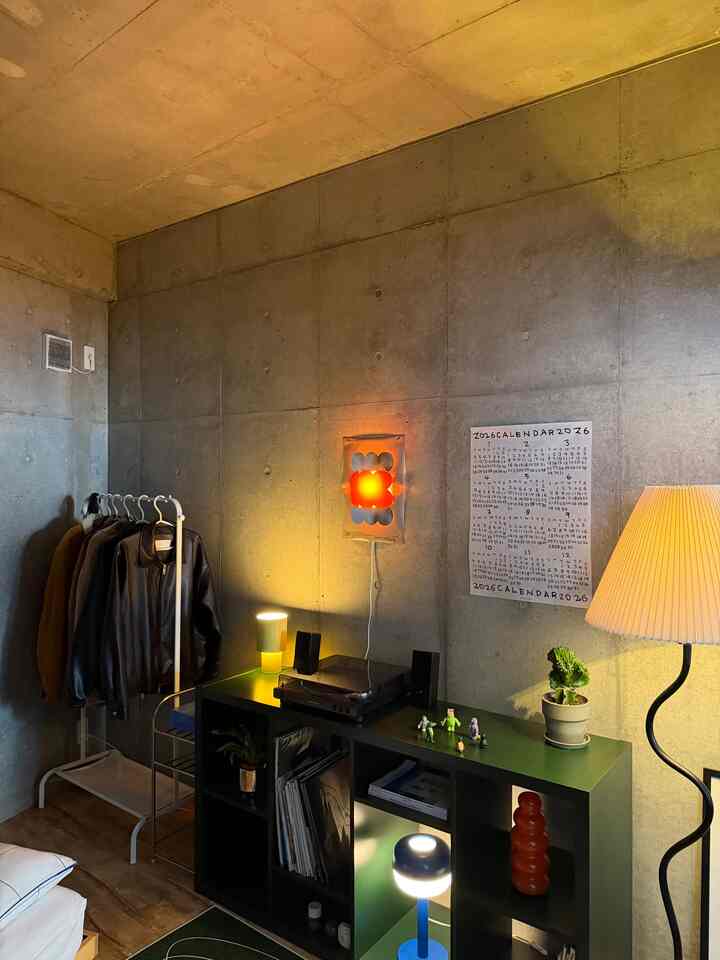 Natural-toned studio apartment with industrial concrete walls featuring a floor lamp, storage unit, and record player in a minimalist setting
