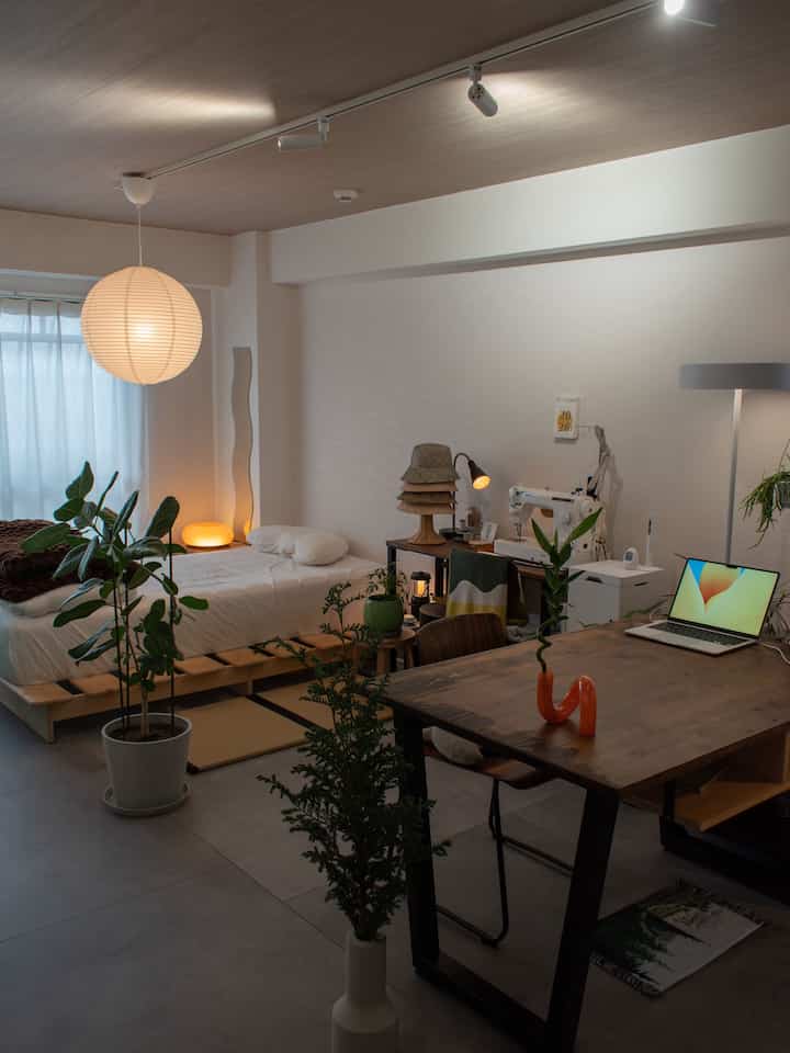 ナチュラルカラーと白を基調としたワンルームの寝室、木製家具とペーパーシェード照明が配置された落ち着いたホームオフィス空間