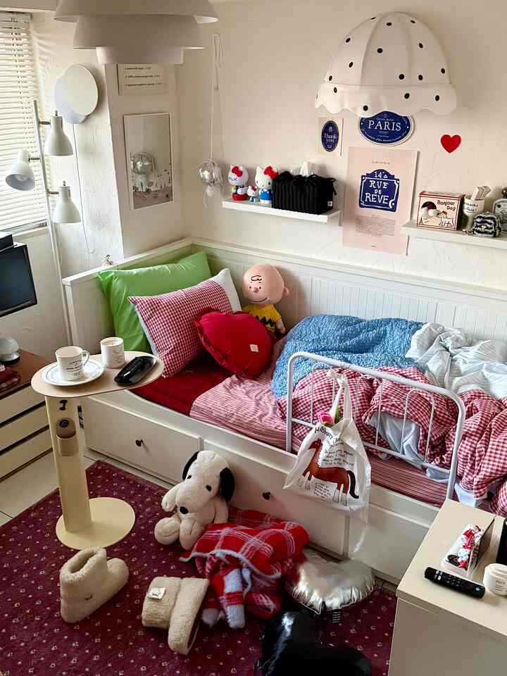 A cozy small bedroom in white and red tones featuring a sofa bed with checkered patterns, cushions, and cafe-style decor