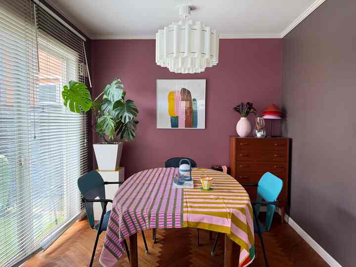 ピンクとブラウンの壁に囲まれたリビングダイニング、カラフルなテーブルクロスをかけた中央のダイニングテーブルと椅子、ペンダントライトと大きな緑の植物が配置された雰囲気のあるインテリア
