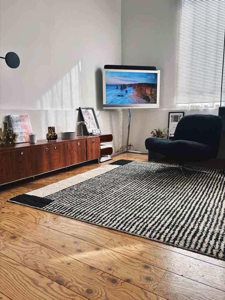 ホワイトの壁と木目調のテレビ台、ブラックのアームチェアとパターンカーペットが配置されたミッドセンチュリーモダン風のリビングスペース