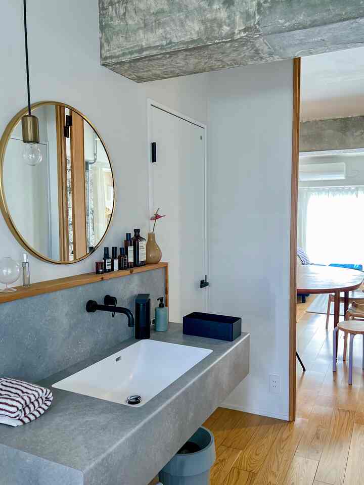 白い壁とグレーの洗面台があるバスルーム、木目調の床と丸い金色の鏡が配置された雰囲気のあるモダンな空間