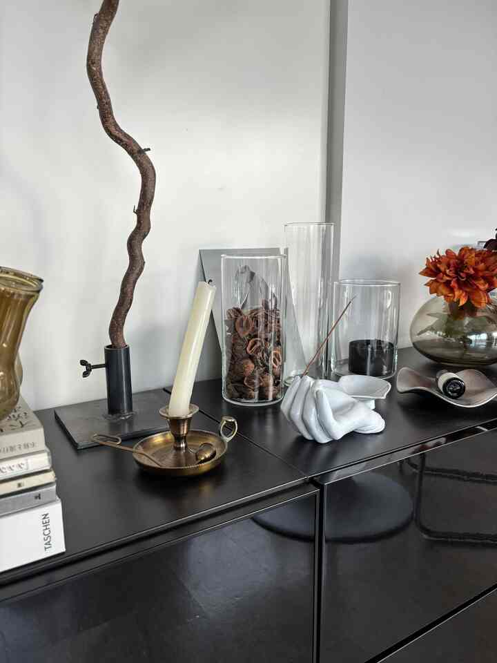 黒い金属キャビネットの上にガラスの花瓶とキャンドルホルダー、自然素材の装飾が置かれたミニマルでインダストリアルなリビングスペース