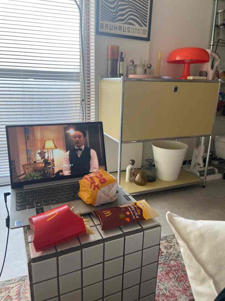 明るい黄色のサイドボードと赤いテーブルランプがある一人暮らしのリビング、ノートパソコンとマクドナルドの食べ物が置かれたタイルのテーブルがあるシンプルモダンな空間