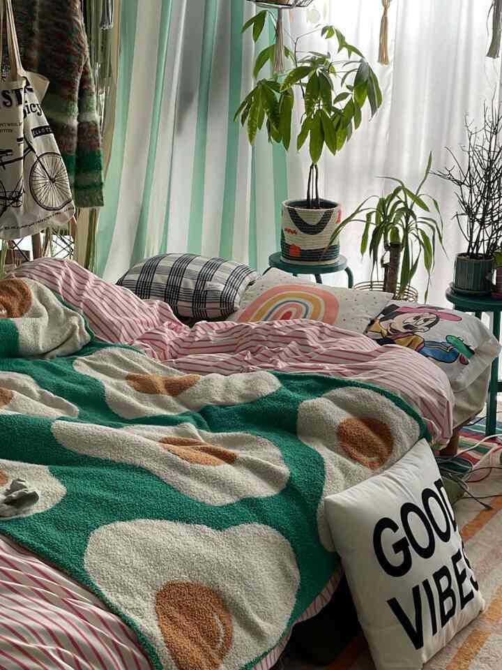 Cozy bedroom with green and cream floral throw blanket, plants on stands, and striped curtains in a single household space