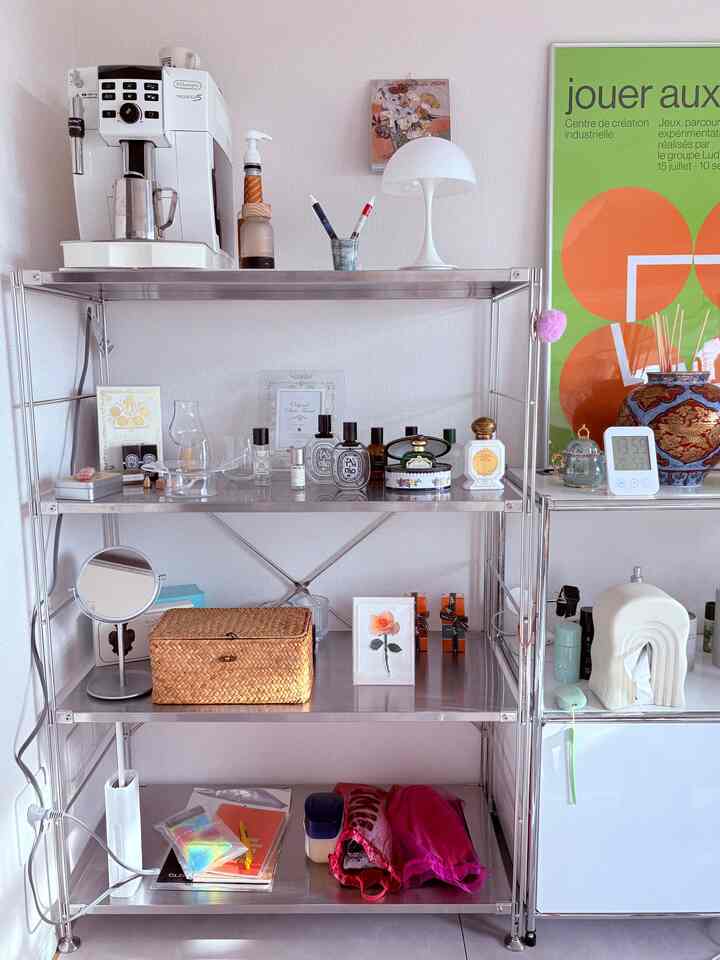 Light gray and white toned silver steel rack featuring various perfumes, a coffee machine, and a table lamp in a neat interior space