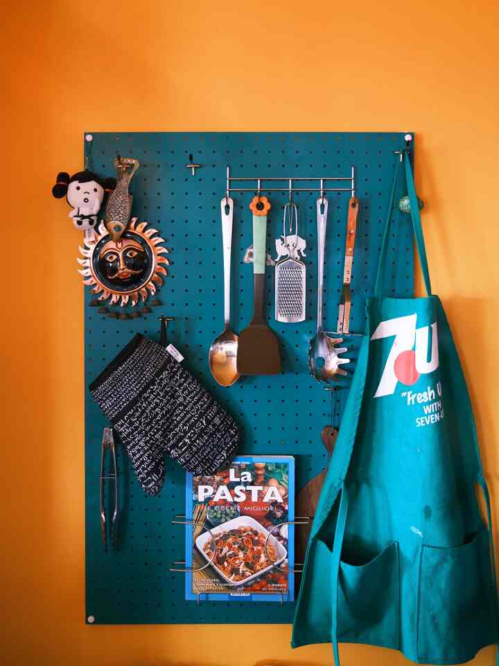 オレンジの壁とターコイズのペグボードが印象的なキッチン、調理器具とエプロンが配置された雰囲気のあるインテリア