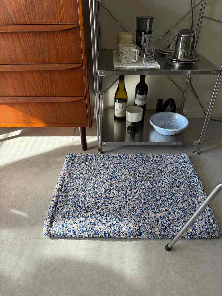 ブラウンの木製チェストとシルバーのメタルシェルフ、青とベージュが混ざったバスマットが配置されたシンプルな室内空間