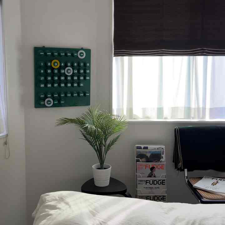 白い壁と自然光が調和する小さな部屋、緑の植物を置いたスツールと椅子、壁に掛けられた永久カレンダーがあるシンプルなインテリア