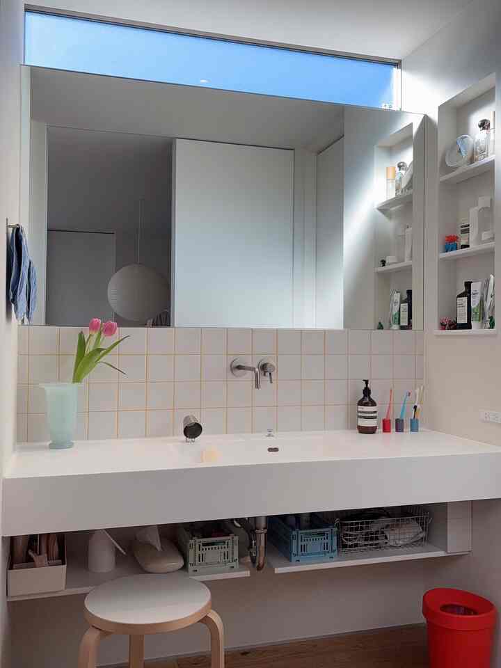 White-toned bathroom featuring a large mirror, narrow elongated space with a stool and organized storage under the sink