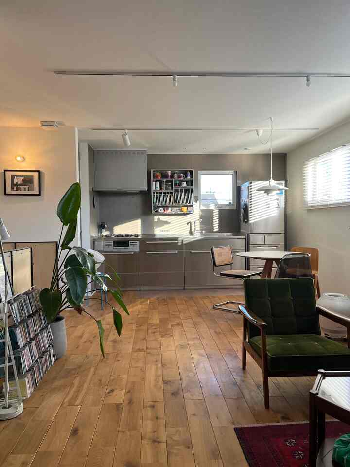 暖かい木目調の床とステンレスキッチンがあるミッドセンチュリーモダンなリビング兼ダイニング空間、グリーンのアームチェアと丸いダイニングテーブルが配置された居心地の良いインテリア