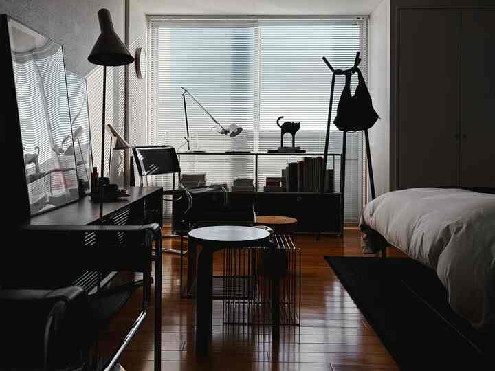 白とウッドトーンが調和する寝室兼ホームオフィス、窓にブラインドがあり、デスクやベッド、コートラックが配置された雰囲気のある空間