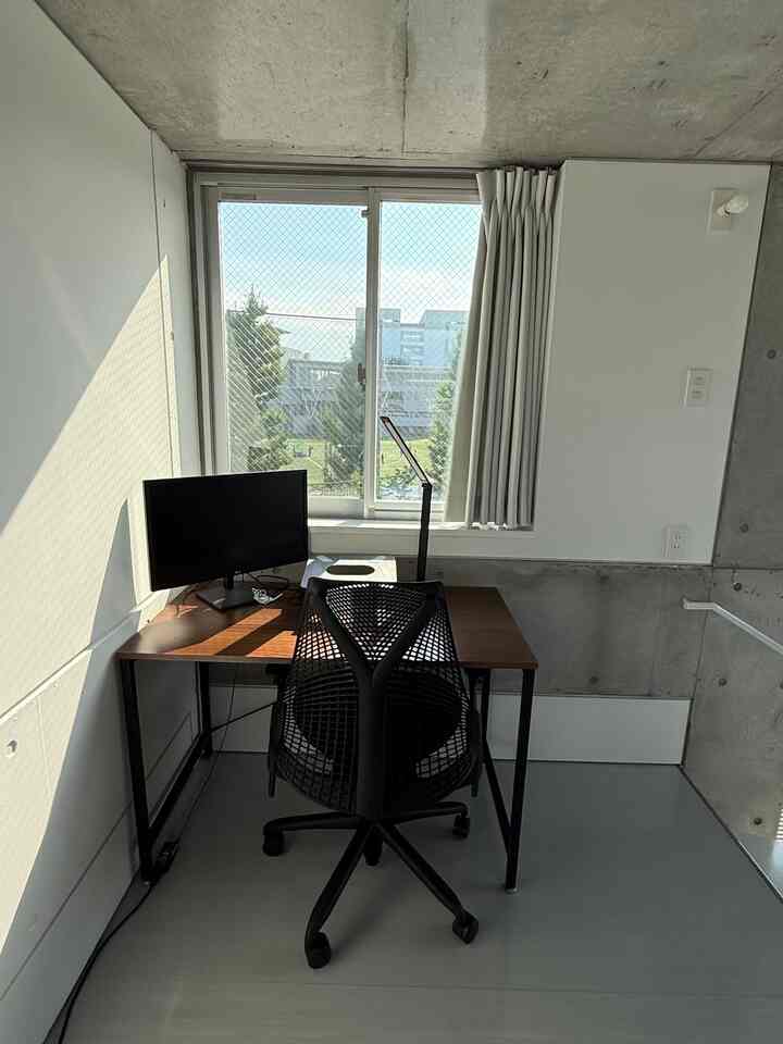 白い壁とコンクリート壁が調和する小さなホームオフィス、茶色のデスクと黒いオフィスチェアが配置された印象的な空間。