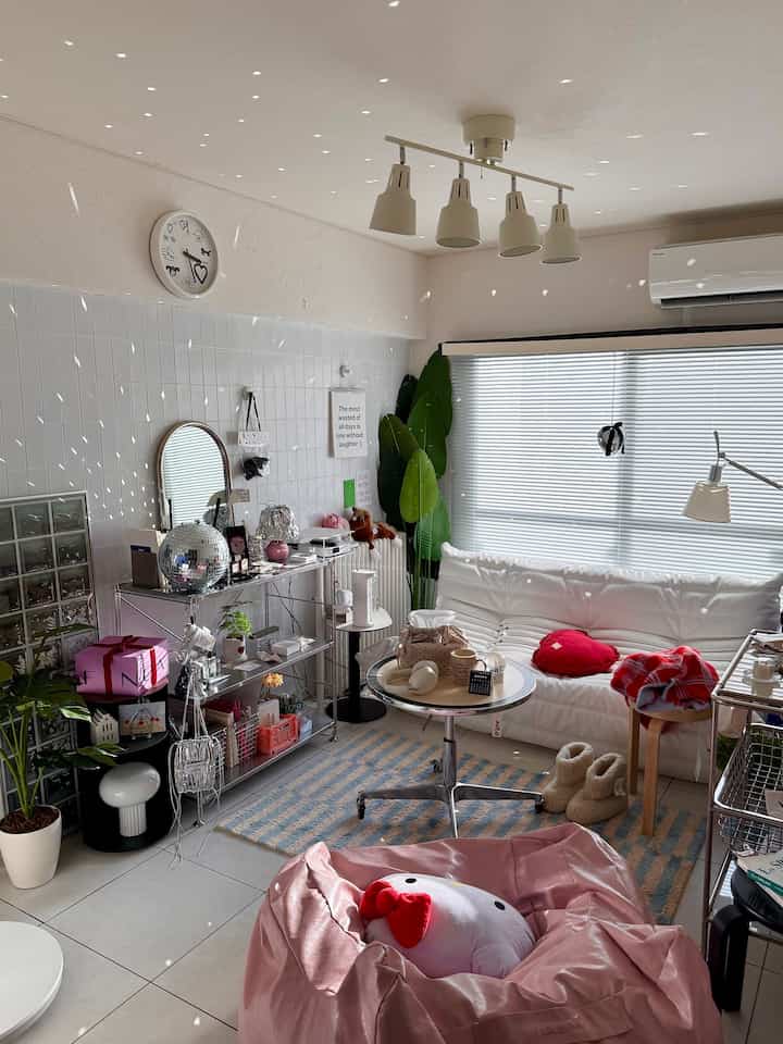 White and pink toned living room featuring a white sofa, pink bean bag, blinds-covered window, and green plants creating a cozy atmosphere