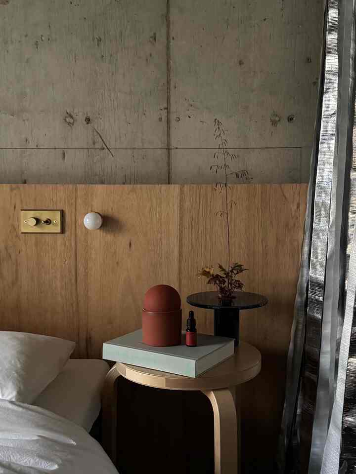グレーのコンクリート壁と淡い木目調のヘッドボードがある寝室、ナイトテーブルに丸いキャンドルとアロマオイルが配置された雰囲気のあるインテリア