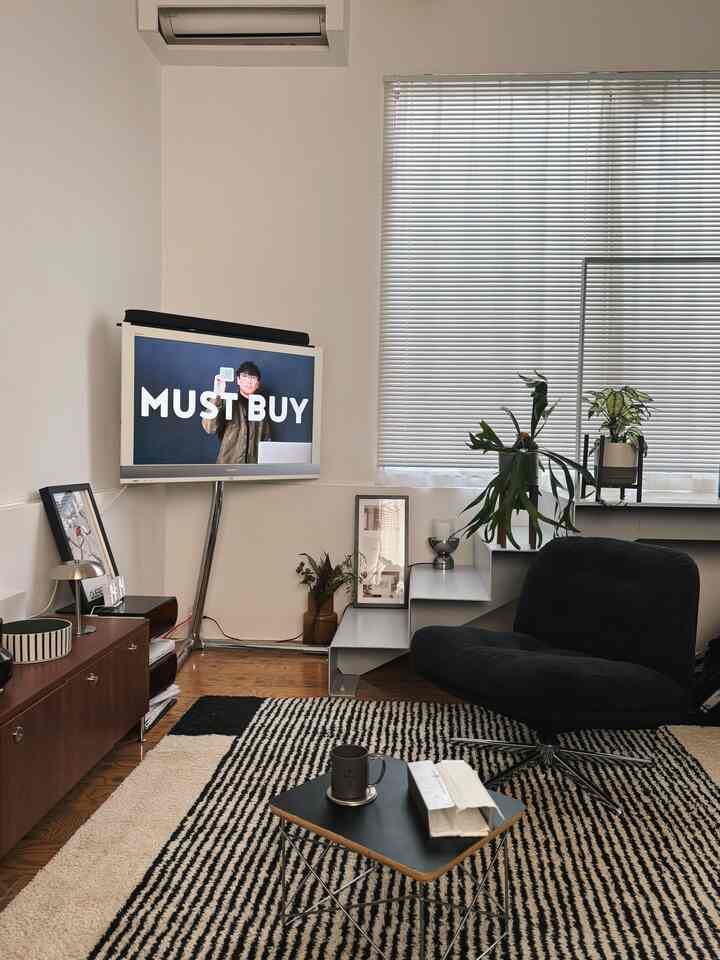 ブラウンの木目調床と黒いアームチェアを配したミッドセンチュリーモダンなリビング、柄のある大きなラグと壁掛けテレビが配置されたすっきりとした空間