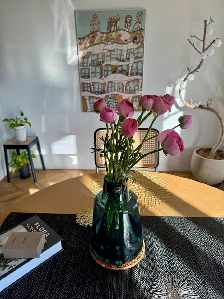 A cafe-style dining room with white walls and wood tone table, featuring a central pink vase with flowers and plants, creating a natural and simple atmosphere