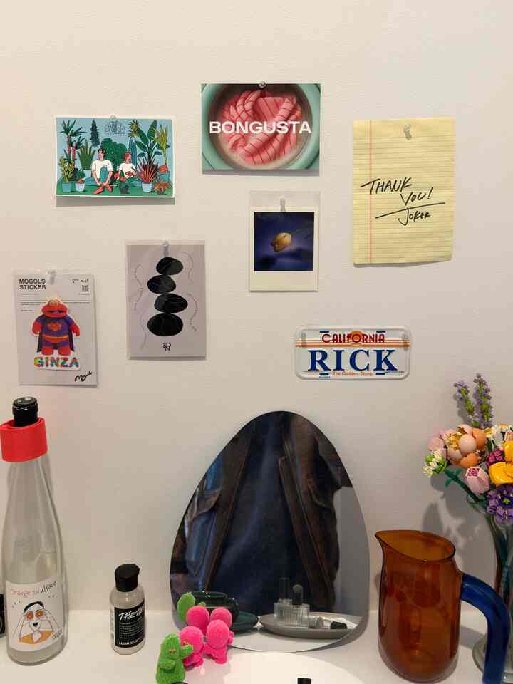 明るいベージュの壁に様々なアートやメモが貼られ、三角形の鏡と小物が置かれたミニマルでトレンディな空間