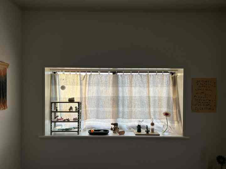 A studio apartment window featuring white walls and natural-tone curtains, decorated with various objets and a vase creating a cozy atmosphere