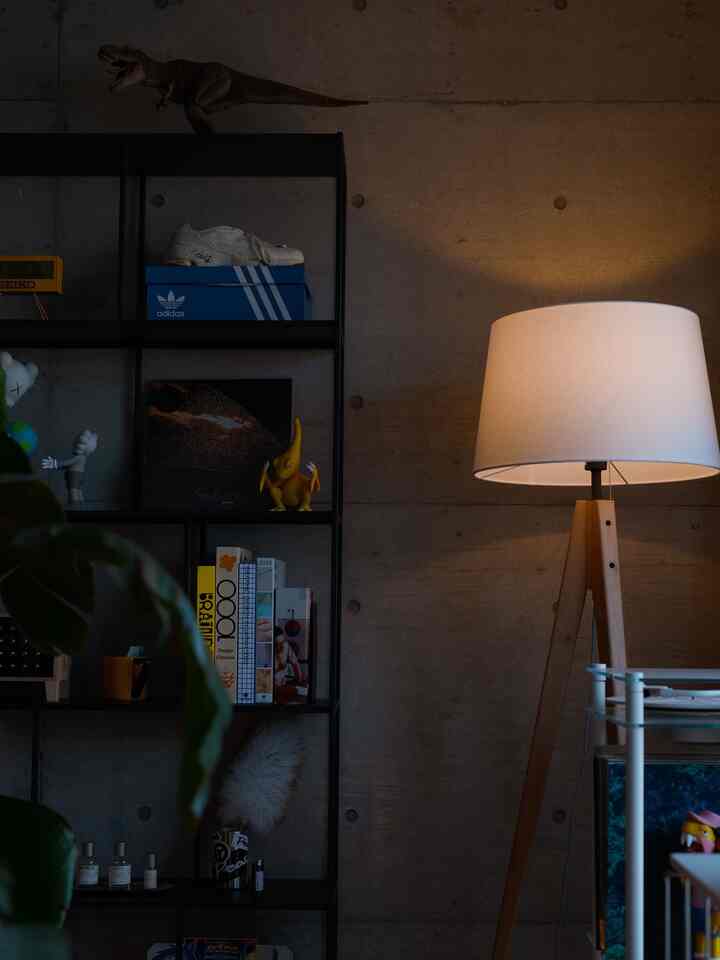 グレーのコンクリート壁と黒の棚、木製トリポッドスタンドの白いフロアランプが配置されたモダンな1LDKのリビング空間