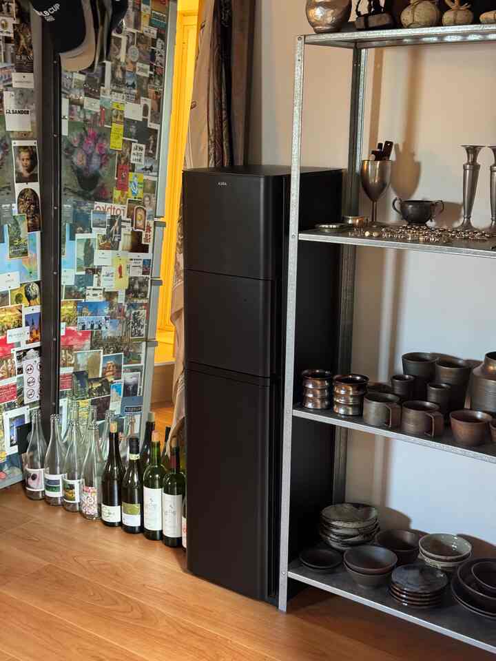 ブラウンの木製床のある1LDKの一人暮らしキッチンスペース、黒のウォーターサーバーとキッチン用品を置いたメタル棚が配置されている