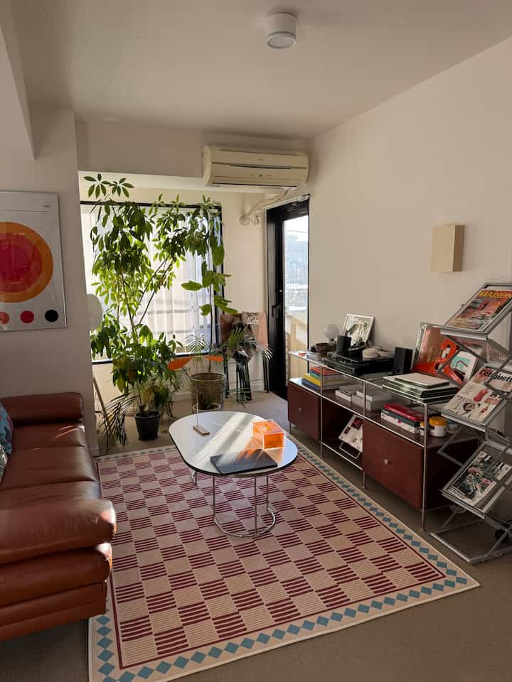 Warm natural-tone living room featuring brown leather sofa, red checkered rug, and green plants creating a cozy atmosphere