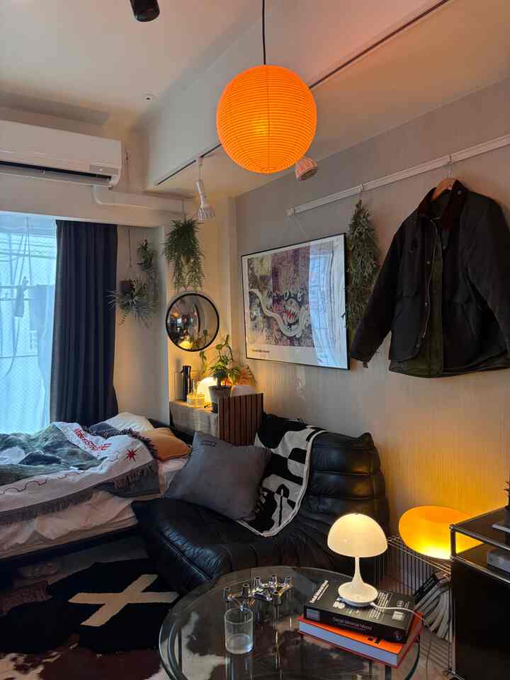 暖かい光と自然の要素がある8畳の1K一人暮らしのリビング、黒いレザーソファとベッドが調和した居心地の良い空間