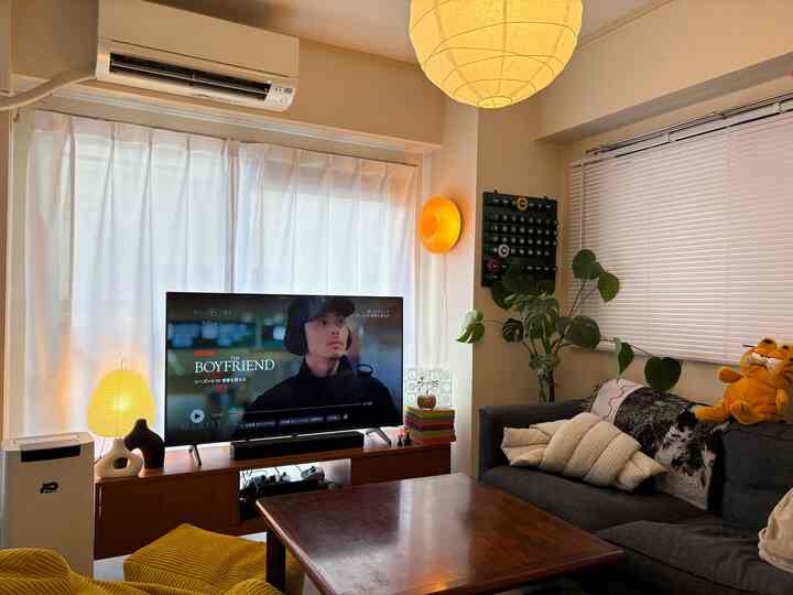 アイボリーとブラウンのトーンの12畳リビング、ミディアムサイズのソファと木製テレビ台、カーテンと植物が配置された雰囲気のある空間