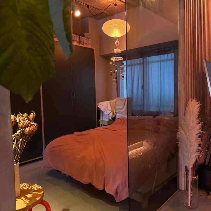 ベージュの壁とカーテンがあるワンルーム寝室、オレンジのベッドカバーと丸いペンダントライト、ナチュラルなインテリアが配置された落ち着いた空間