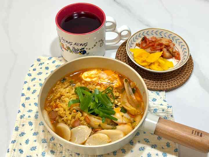 白いテーブルの上にベージュ色の鍋とラーメン、隣に小皿と紅茶が置かれた雰囲気のあるキッチン空間