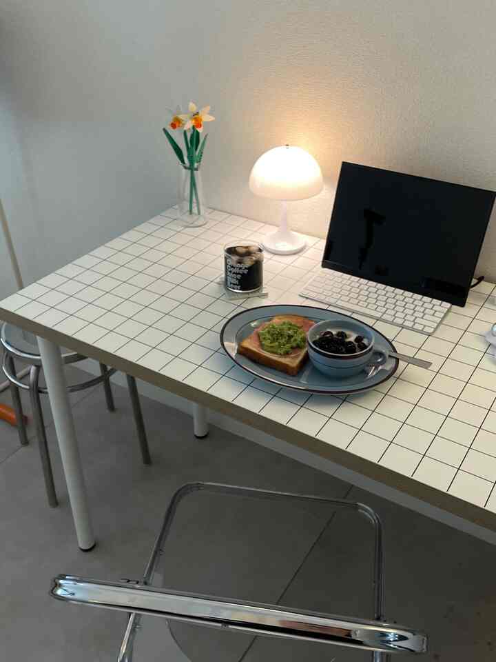 白いタイルのテーブルに青い皿と朝食が置かれ、透明な椅子、小さなランプ、タブレットがあるミニマルなキッチンスペース