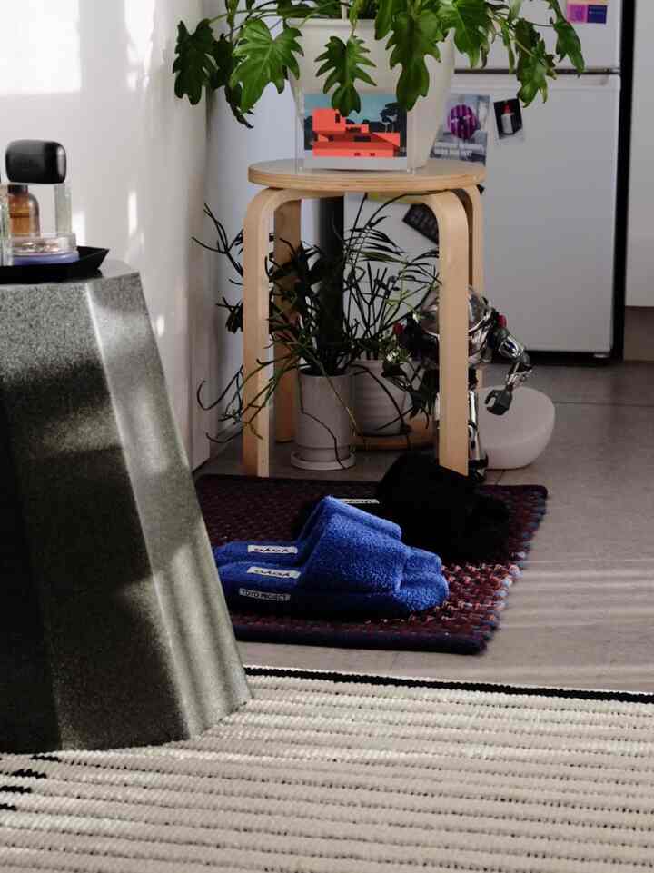 ナチュラルカラーのワンルームリビング、木製スツールと青いスリッパが置かれたラグ、植物が配置された雰囲気のあるインテリア