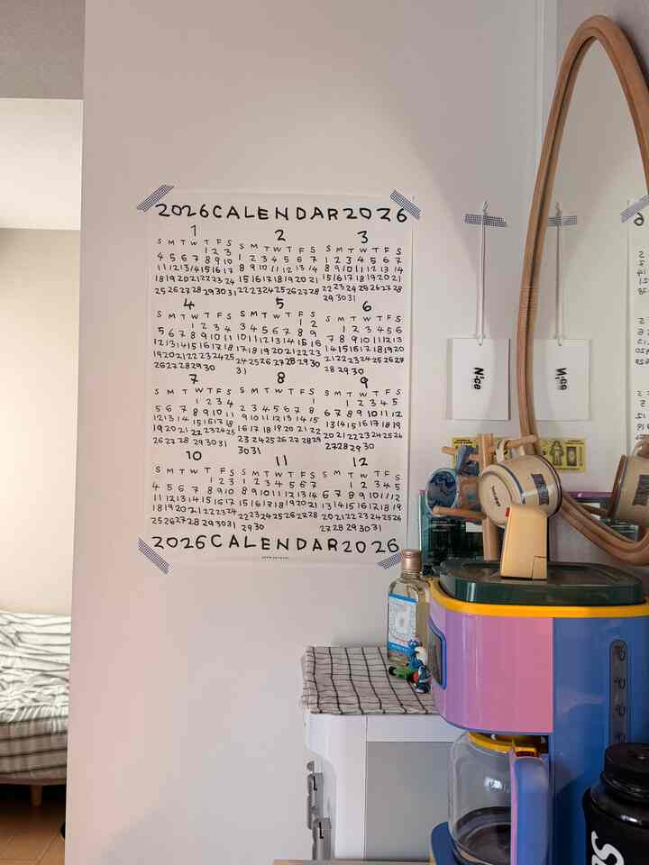 A 6-tatami-sized space with white walls and a calendar on the wall, featuring a colorful coffee maker and wooden framed mirror in a tidy home cafe setting