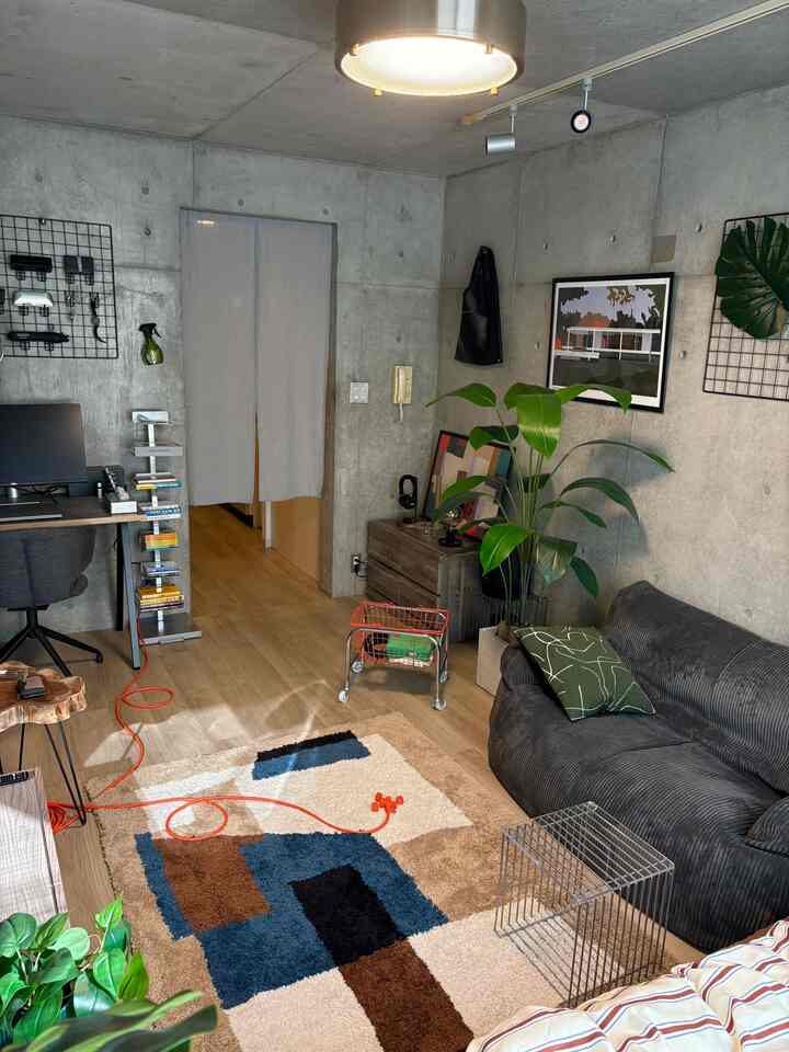 グレーのコンクリート壁とブラウンの木目調の床があるモダンな1Kスタジオ、ソファとデスク、ジオメトリックラグが配置された印象的な空間