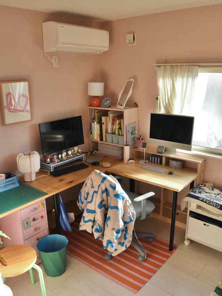 柔らかなピンク色の壁と木製デスクが配置された、二台のコンピューターとオフィスチェアがある温かみのあるホームオフィス空間。