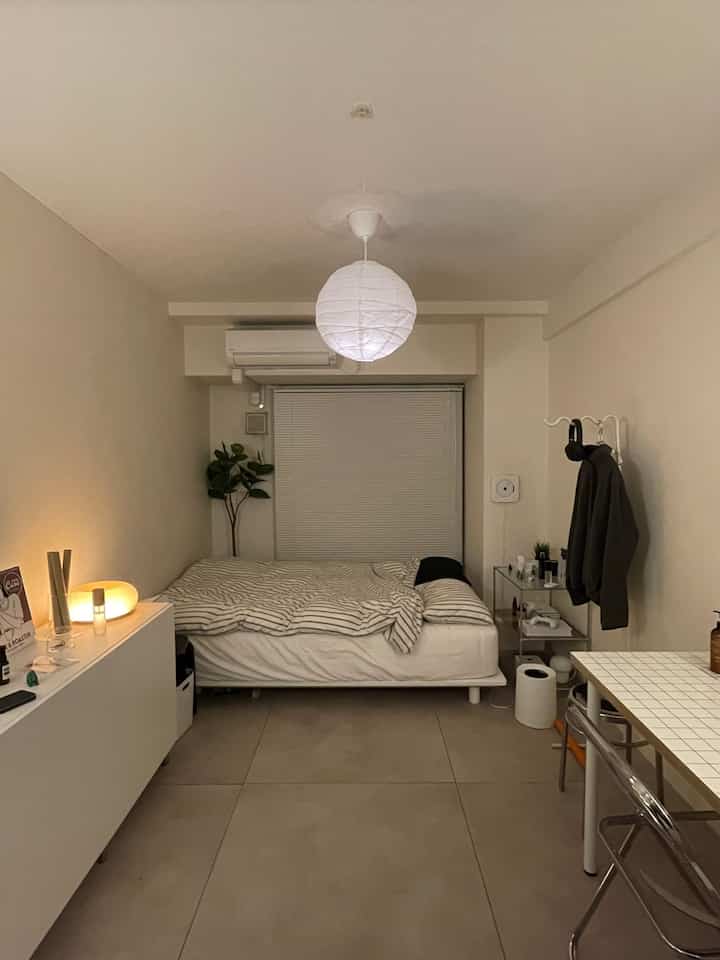 Natural beige-toned 1K bedroom featuring a white bed with striped bedding centrally, table lamp on side, and clothes rack on right wall