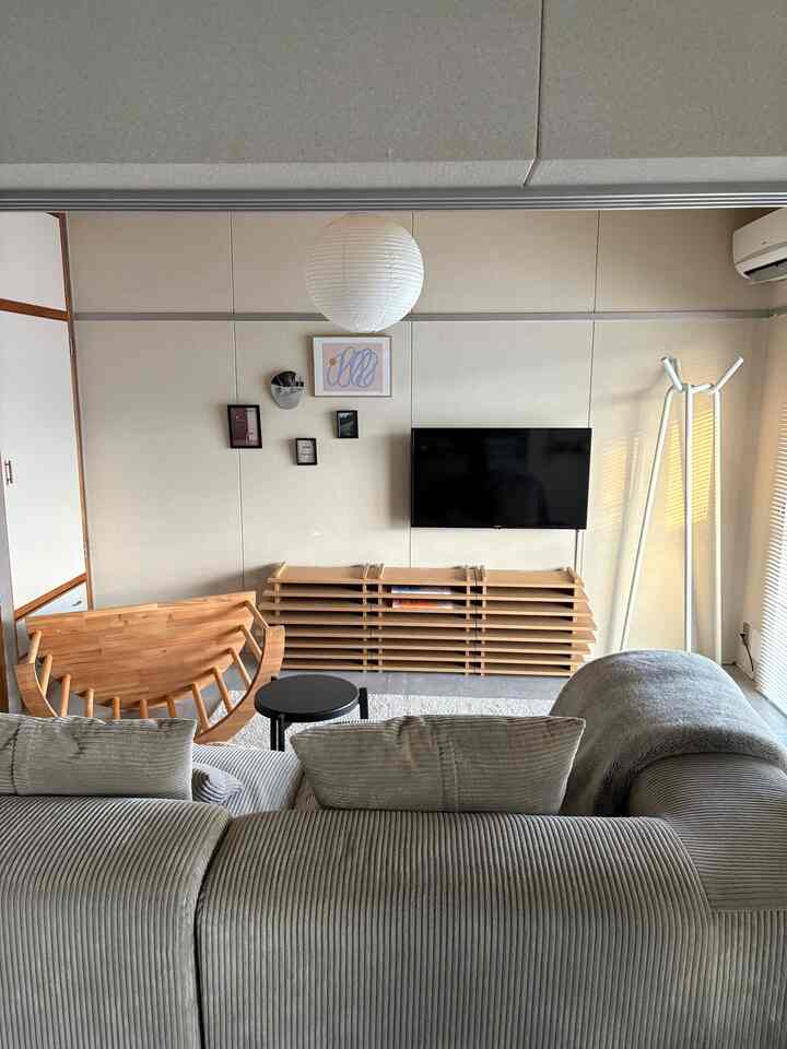 A modern living room with bright beige walls, gray fabric sofa, natural wood TV console, and white pendant light