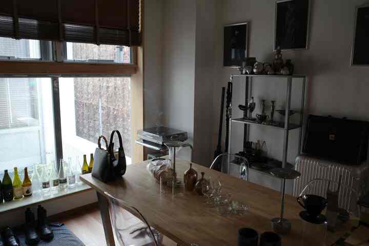 ナチュラルな色調の1LDKリビング、木製ダイニングテーブルと透明な椅子、金属製の棚と窓のブラインドが配された雰囲気のある空間