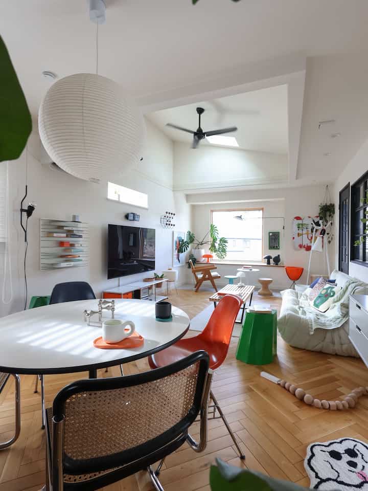 Bright mid-century modern living room with white walls and ceiling, featuring orange and green accents, sofas and various stools arranged in an open layout