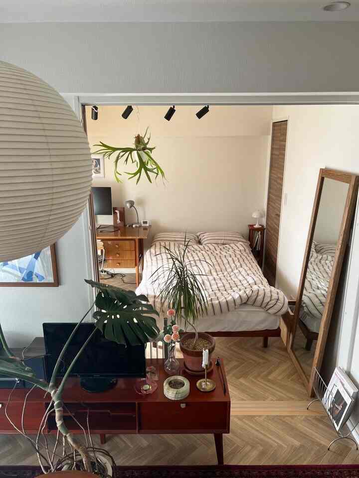 ナチュラルな色調のワンルーム寝室、ミッドセンチュリーモダンのベッドとデスク、多数の観葉植物が配置された雰囲気のあるインテリア