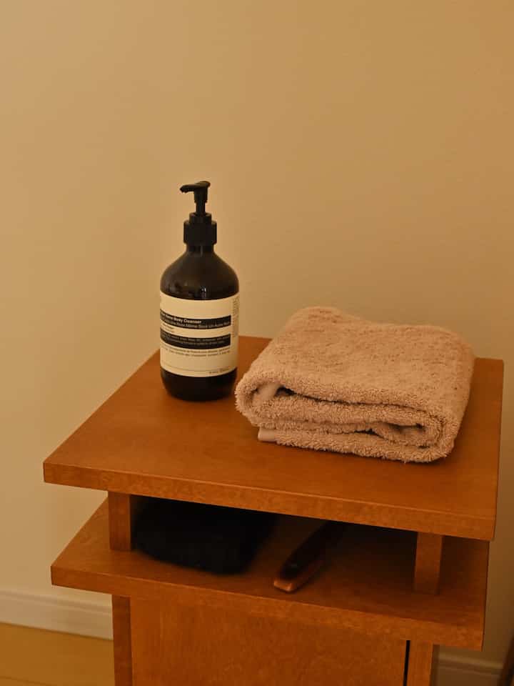 ブラウンの木目調サイドテーブルにベージュのタオルと黒のボディクレンザーが置かれたナチュラルなバスルームの一角