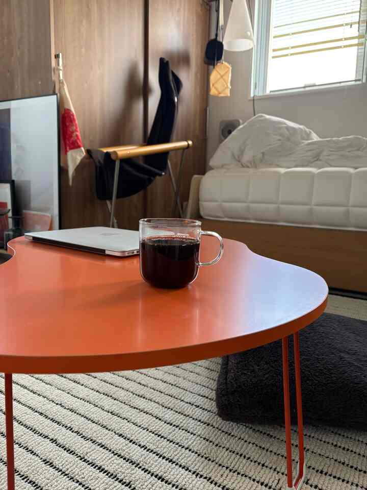Studio apartment living room with wood tone walls and white bed, featuring orange folding table and black armchair in a cozy setting