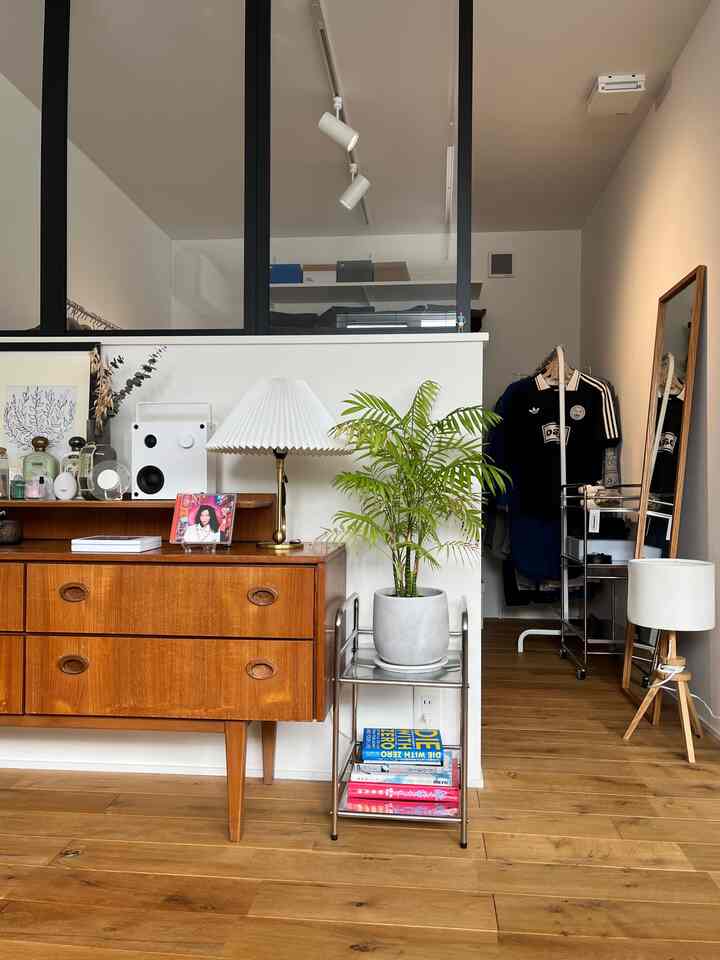 白い壁と木目調の床が調和するミッドセンチュリーモダンな空間、ヴィンテージ家具とスチールラックに植物と照明が配置された雰囲気のあるインテリア